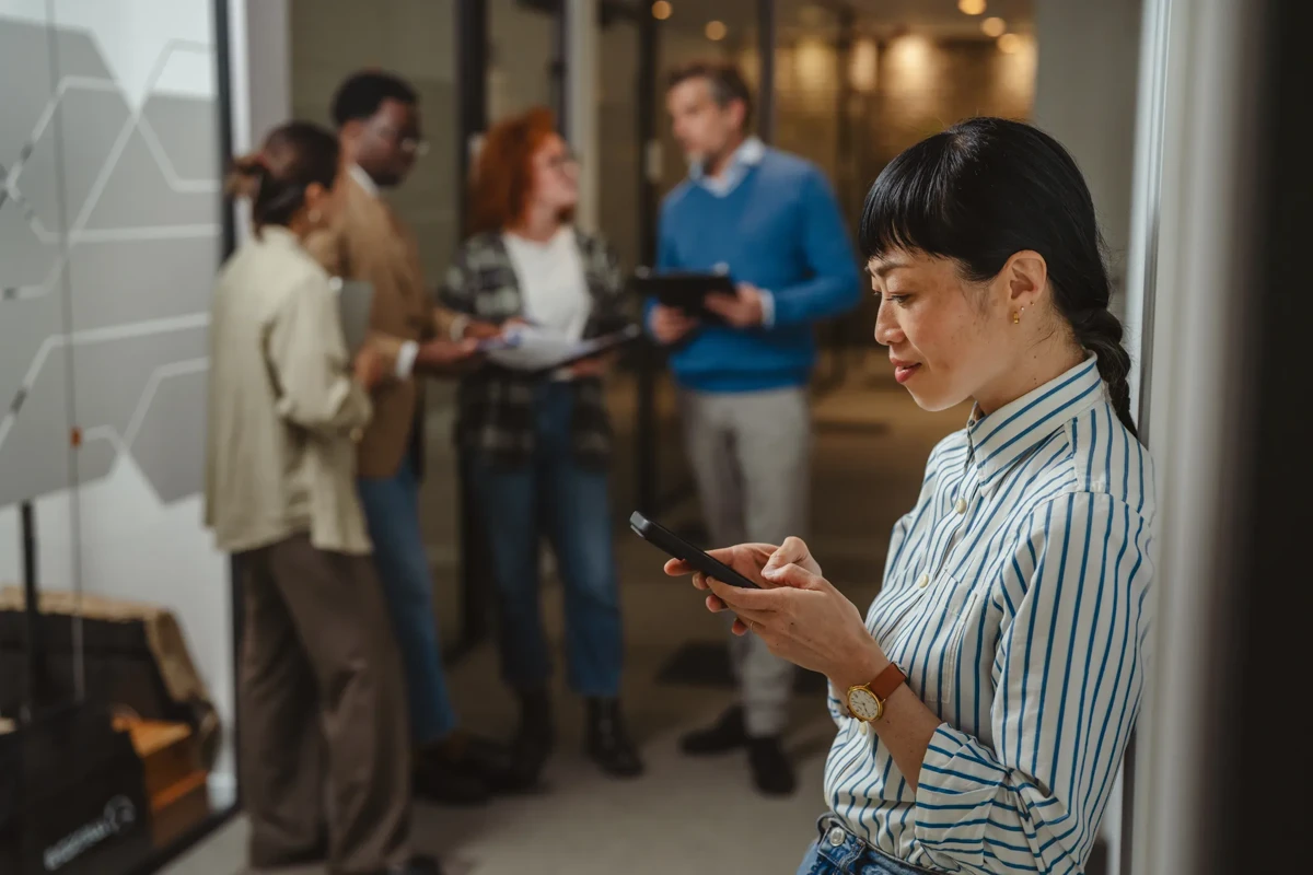 Asian businesswoman checking messages on her mobile phone while leaning against a wall, with a diverse group of busy colleagues collaborating in the background of their modern office